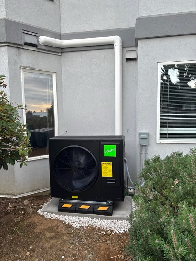 Black Jetson Air 3-ton central ducted heat pump condenser installed against grey stucco exterior in Abbotsford, with white PVC line-set conduit running up the wall and decorative white gravel surroundin the unit.
