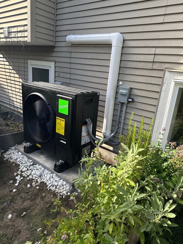 Black Jetson Air heat pump condenser against grey vinyl siding with PVC conduit routed to an electrical disconnect box, garden shrubs and flowering plants in the foreground, Abbotsford.