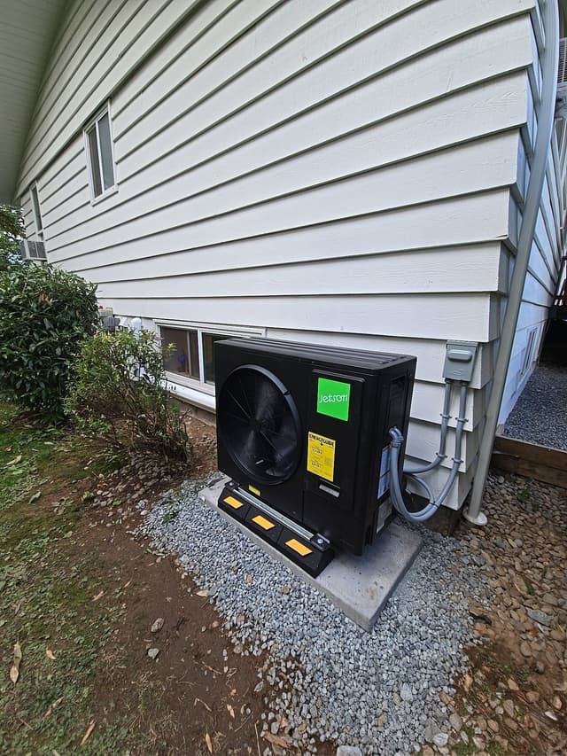 Black Jetson Air 3-ton heat pump condenser along the side of a grey vinyl-sided house in Abbotsford, with grey conduit and refrigerant lines running neatly up to an electrical disconnect box.