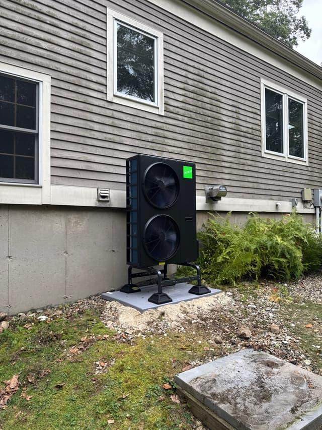 A black Jetson Air 4-ton heat pump installed along the side of a gray-sided house in Andover, with ferns and shrubs nearby.