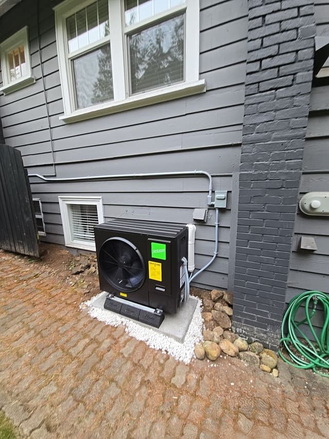 Jetson Air central ducted heat pump condenser installed against the gray lap-siding and brick exterior of an Anmore home, with cobblestone pavers and a garden bed border visible in the foreground.