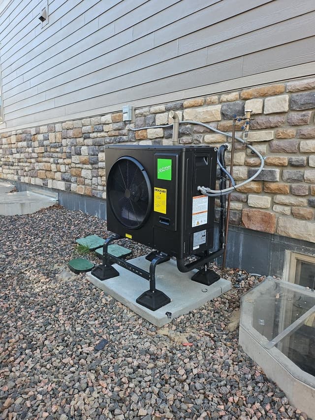 Black Jetson Air 3-ton heat pump mounted on a raised metal stand on a concrete pad against the stone and lap siding exterior of a home in Aurora, CO, with refrigerant lines and conduit running neatly up the wall.