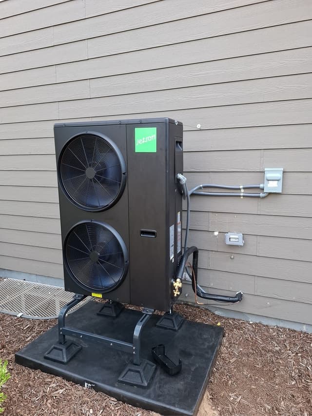 Jetson Air 5-ton dual-fan heat pump condenser on a black pad in a mulched bed alongside a gray-sided house in Aurora, CO, with refrigerant lines and conduit running to a disconnect box and electrical panel on the wall.