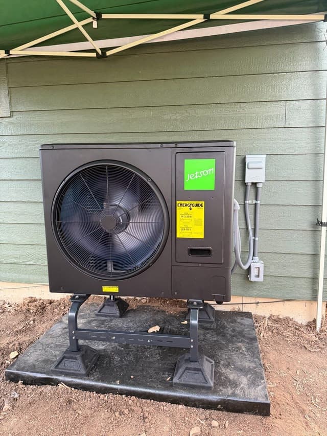 Jetson Air 3-ton heat pump mounted on a riser stand on bare dirt, with electrical conduit and disconnect box neatly installed on the green lap-siding wall beside it, Aurora, CO.