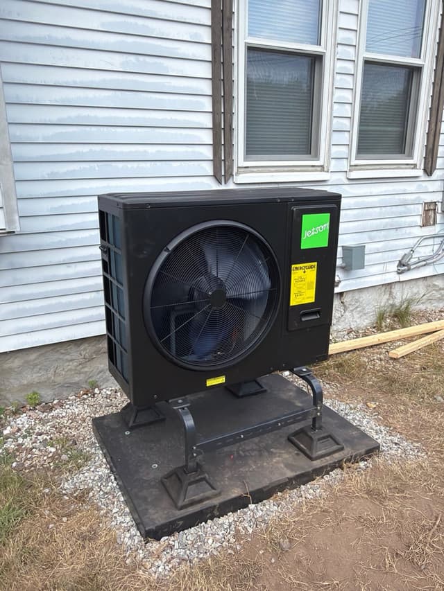 Jetson Air 3-ton heat pump mounted on a black metal stand over a gravel bed against grey vinyl siding of a home in Avon, MA.