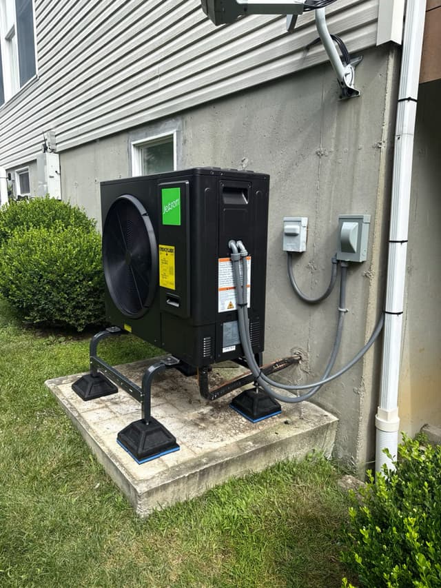 Black Jetson Air 3-ton heat pump installed against a gray stucco foundation with vinyl siding above, refrigerant lines and two electrical disconnect boxes on the wall, green shrubs to the left, Baldwin Place NY.
