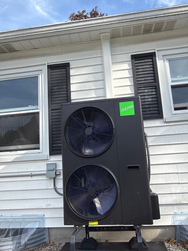 Jetson Air 4-ton heat pump condenser mounted on a metal stand against white vinyl siding of a home in Bellingham, MA, with electrical disconnect box and refrigerant line covers visible.
