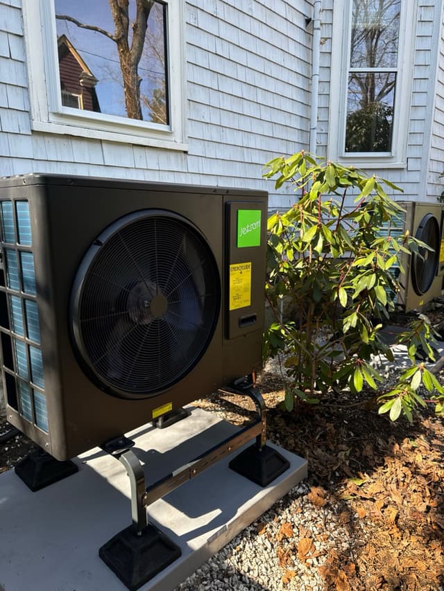 Two Jetson Air heat pump condensers installed on a concrete pad with rubber isolation feet against gray shingle siding of a home in Belmont, MA.