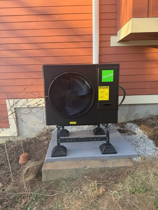 A 3-ton Jetson Air central ducted heat pump mounted on a metal riser stand on a concrete pad along the red-sided exterior of a home in Berlin, MA.