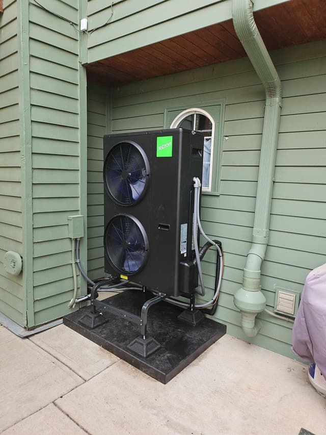 A black Jetson Air 4-ton heat pump on an elevated stand against the green lap-siding exterior of a home in Berthoud, CO, with electrical conduit and refrigerant lines neatly run to a wall-mounted disconnect box.