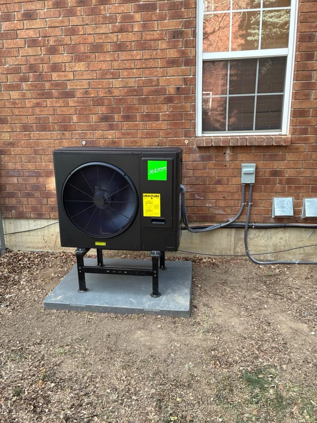 A Jetson Air 3-ton heat pump on a metal stand over a concrete pad against a red-brick house exterior in Berthoud, CO, with black conduit running to a disconnect box along the foundation wall.