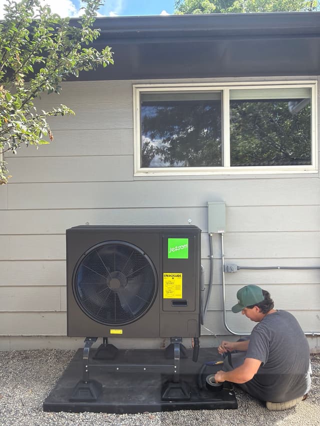 Male technician crouching beside a newly installed black Jetson Air 3-ton central ducted heat pump condenser on the side of a Boulder home, with electrical disconnect and conduit visible on the wall.