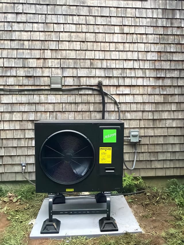 Black Jetson Air 3-ton heat pump condenser mounted against cedar-shingle siding of a home in Bridgewater, with conduit and refrigerant lines running neatly to a disconnect box.