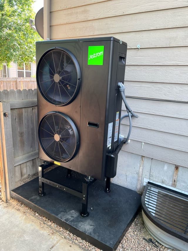Black Jetson Air 4-ton heat pump condenser on a metal stand and rubber pad, mounted between a wooden fence and tan lap siding in the side yard of a Brighton, CO home, with conduit and line set running neatly to a disconnect box.