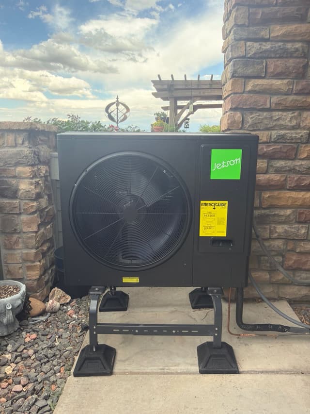 Jetson Air 3-ton central ducted heat pump mounted on a metal stand with rubber feet on a concrete pad, positioned against a stone wall with decorative rock ground cover and a pergola visible in the background, Brighton, CO.
