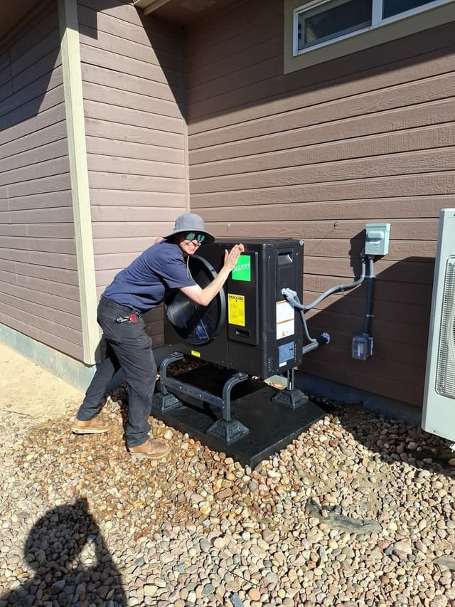 Jetson crew member posing playfully in front of a newly installed black Jetson Air 3-ton heat pump on a metal stand beside a brown lap-siding house in Brighton, CO.