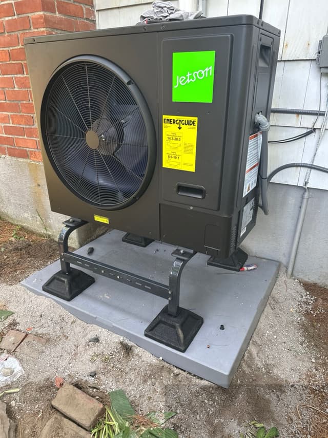 Black Jetson Air 3-ton central ducted heat pump unit mounted between a brick wall and wood-sided house wall in Brockton, MA, with refrigerant lines running into the home.