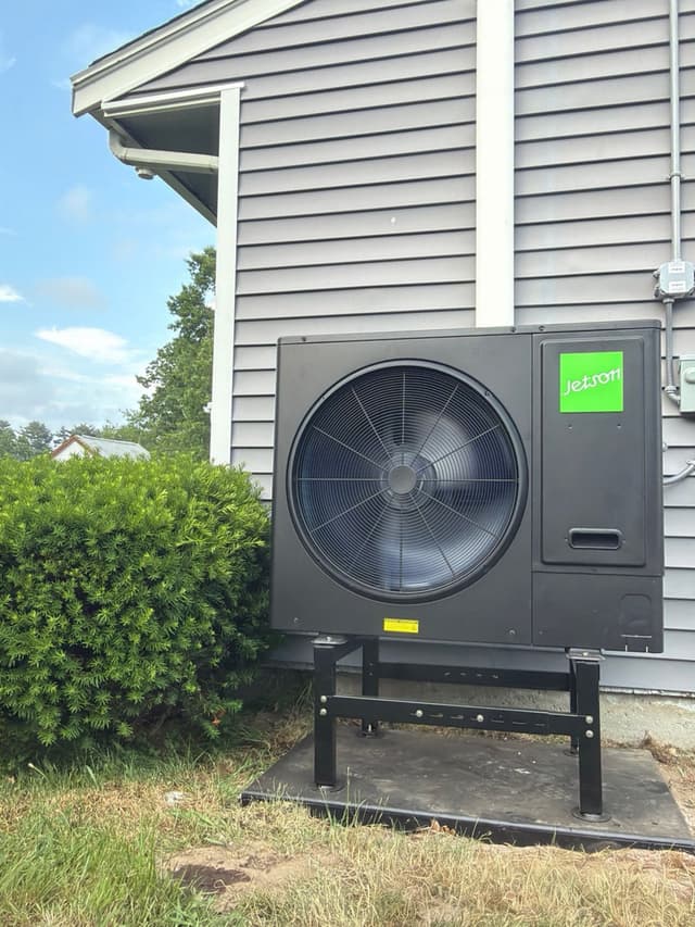 Jetson Air 3-ton outdoor heat pump unit on an elevated metal stand along the side of a gray vinyl-sided house in Brockton, MA, with electrical conduit and disconnect box mounted on the wall beside it.