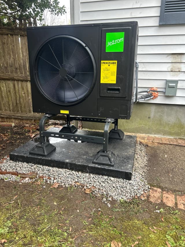 A black Jetson Air 3-ton central ducted heat pump mounted on a raised metal stand in a gravel bed against a white vinyl-sided house in Brockton, MA, with electrical lines running to a disconnect box on the wall.