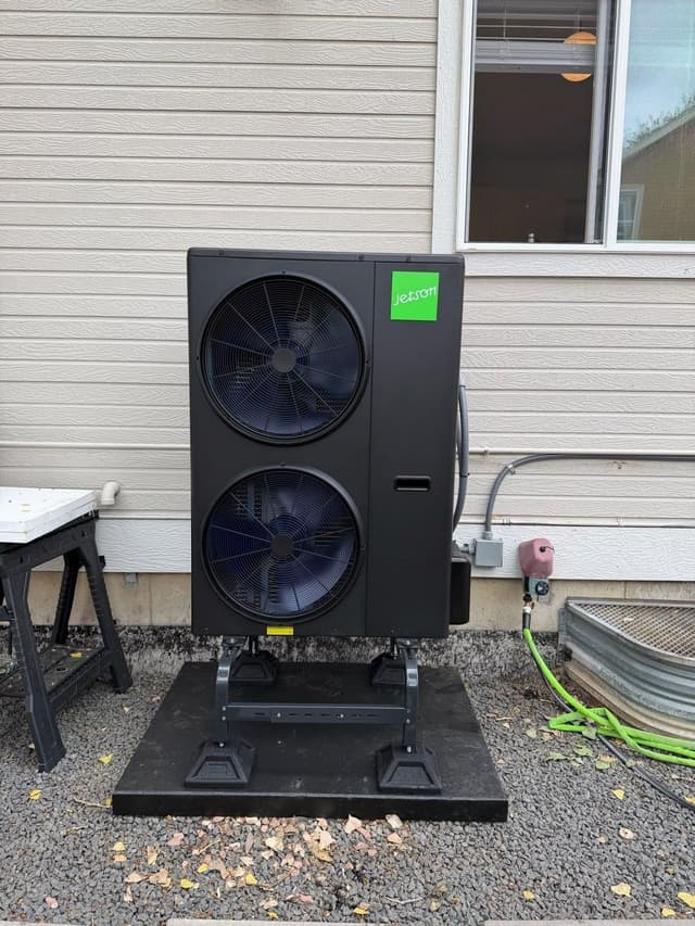 A black Jetson dual fan heat pump condenser on a stand over gravel beside a beige sided house in Broomfield, CO, with electrical conduit running to a disconnect box.