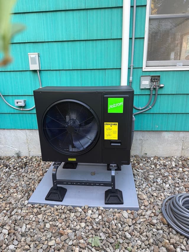 Black Jetson Air 3-ton heat pump condenser installed along the side of a teal-sided house in Burlington, with conduit neatly run along the wall.