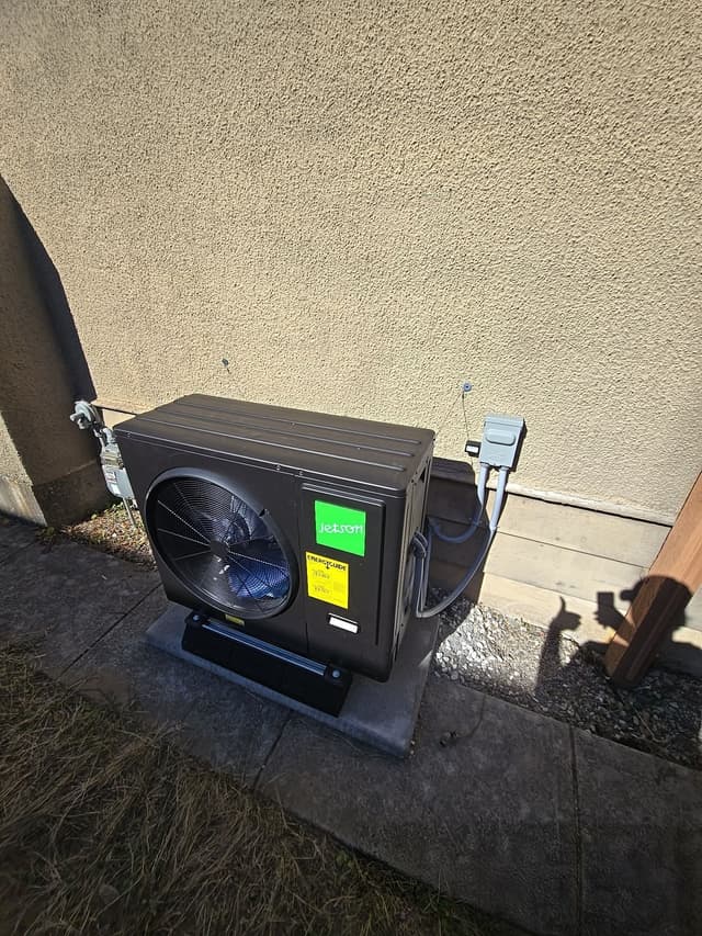 Black Jetson Air 3-ton central ducted heat pump condenser installed against a beige stucco exterior wall at a home in Lougheed, Burnaby, with electrical conduit and disconnect box visible.