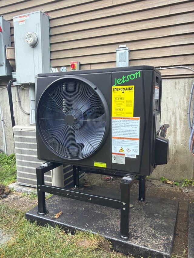 Jetson Air 2-ton heat pump mounted on a black metal stand against tan lap siding on a home in Canton, MA, with an electrical panel and meter visible on the wall beside it.