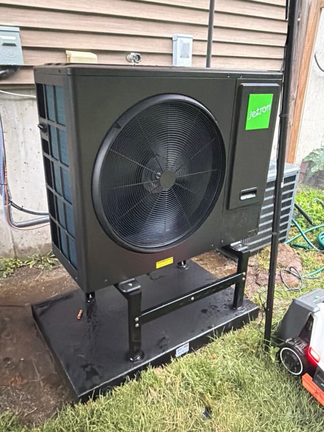 Jetson Air 3-ton heat pump mounted on a black elevated stand beside a house with tan siding in Canton, MA, with wet grass visible at the base after recent rain.