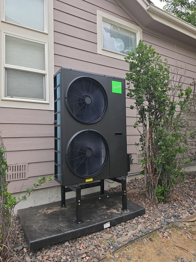 A large black Jetson Air 4-ton heat pump installed along the side of a tan lap-siding house in Castle Pines, with decorative rock ground cover and a leafy shrub to the right.