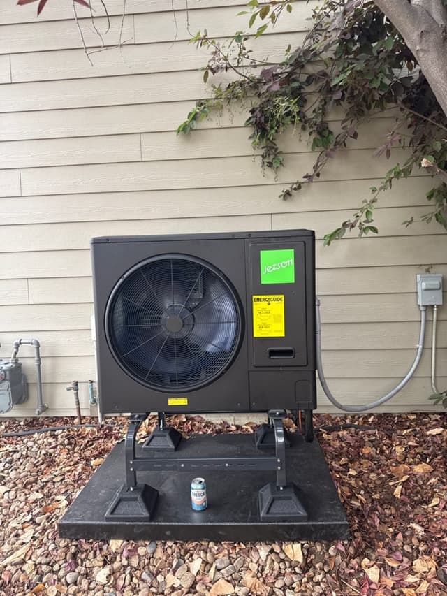 Jetson Air heat pump mounted on a black equipment pad on gravel beside a tan lap-sided house in Centennial, CO, with electrical disconnect and conduit visible on the wall.