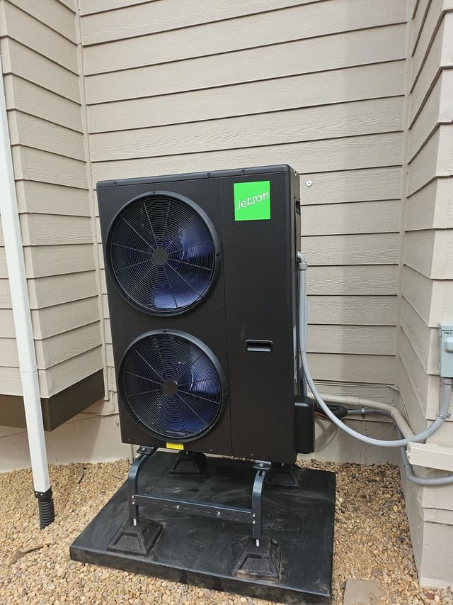 Black Jetson Air 4-ton heat pump mounted on a stand over a gravel bed along the side of a beige lap-siding house in Centennial, CO, with refrigerant lines and conduit running neatly up the wall.
