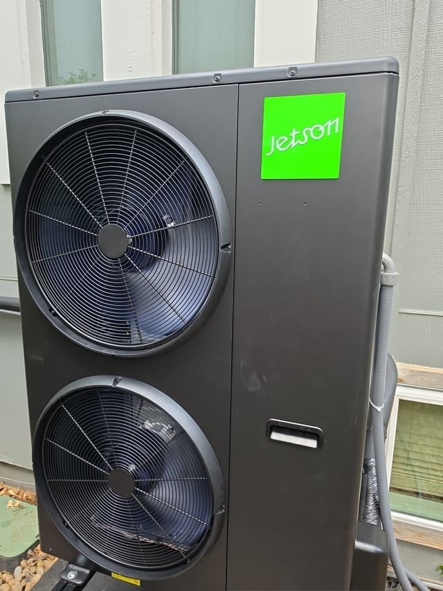 Jetson Air black outdoor heat pump condenser unit installed against gray house siding in Centennial, CO, with two fan grilles and refrigerant lines visible.