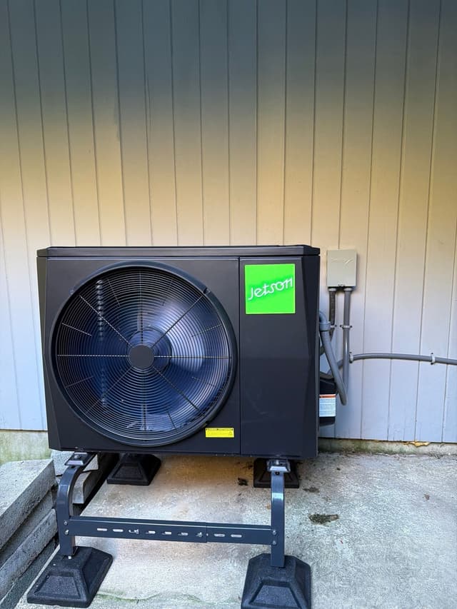 Jetson Air 2-ton central ducted heat pump installed against the side of a light-colored vertical-panel building in Chappaqua, NY, with conduit and refrigerant lines running neatly to a gray disconnect box.