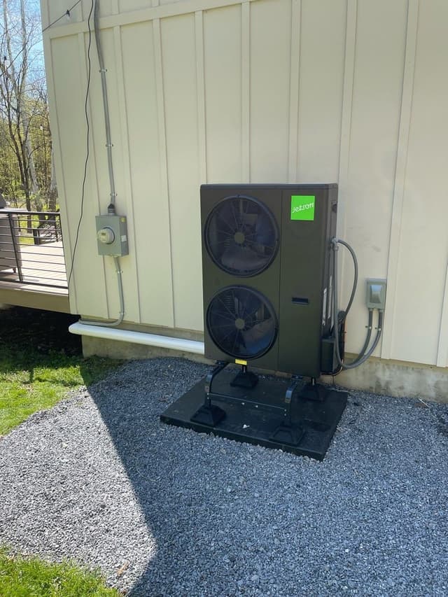 A black Jetson Air 5-ton outdoor heat pump condenser unit mounted on a riser pad over a gravel bed beside a white vertical-board house in Clinton, NY, with refrigerant lines and electrical conduit running neatly up the wall to a disconnect box.