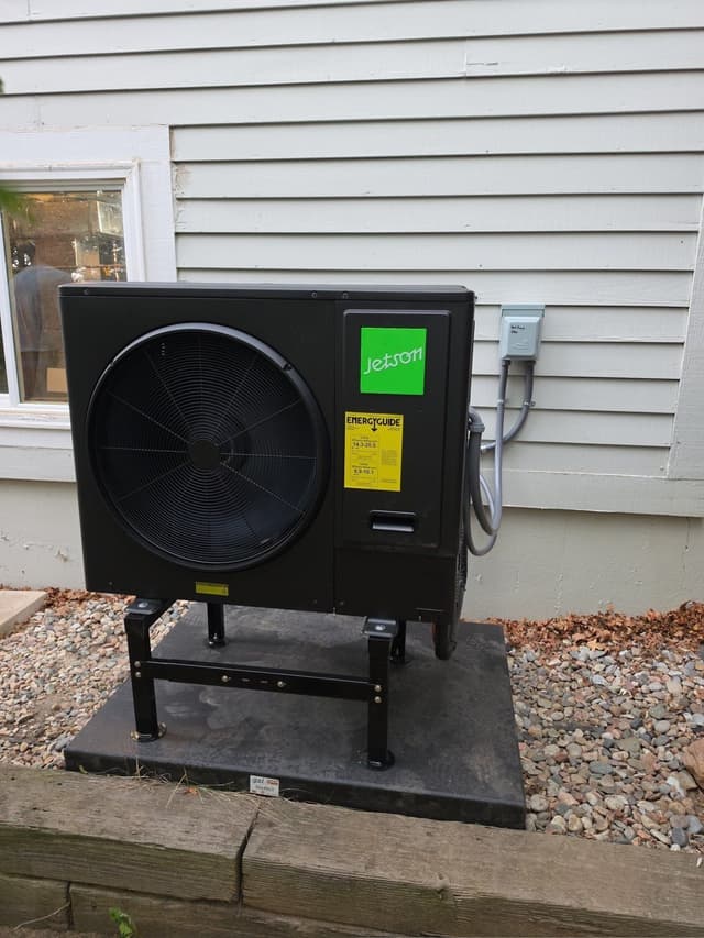 Jetson Air 3-ton central ducted heat pump mounted on a metal stand over a gravel pad against gray vinyl siding, with conduit and disconnect box visible, at a Colorado Springs home.