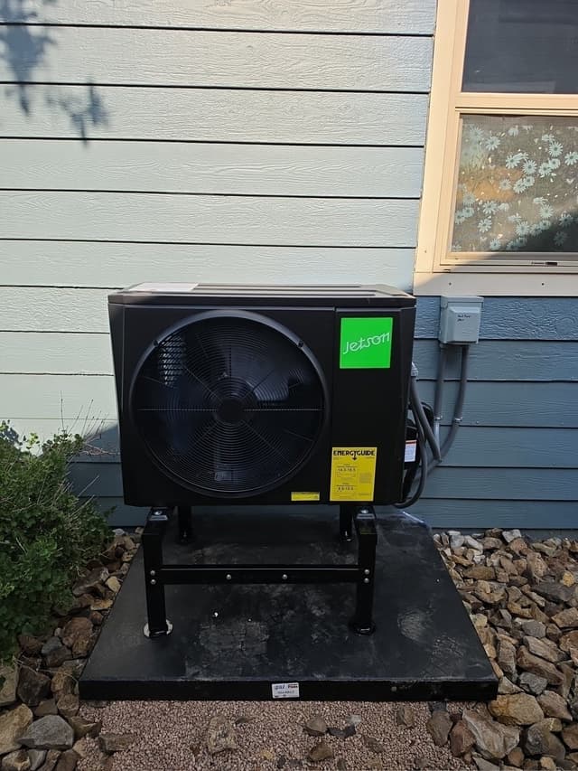 Black Jetson Air heat pump condenser on a metal stand and concrete pad with gravel, mounted against light-blue house siding in Colorado Springs, CO.
