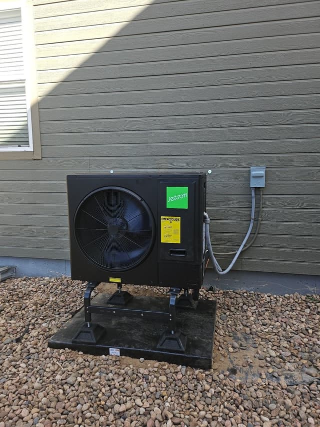 A black Jetson Air 3-ton heat pump mounted on a pad in a gravel bed alongside a gray lap siding house in Commerce City, CO, with conduit running up to a wall-mounted disconnect box.