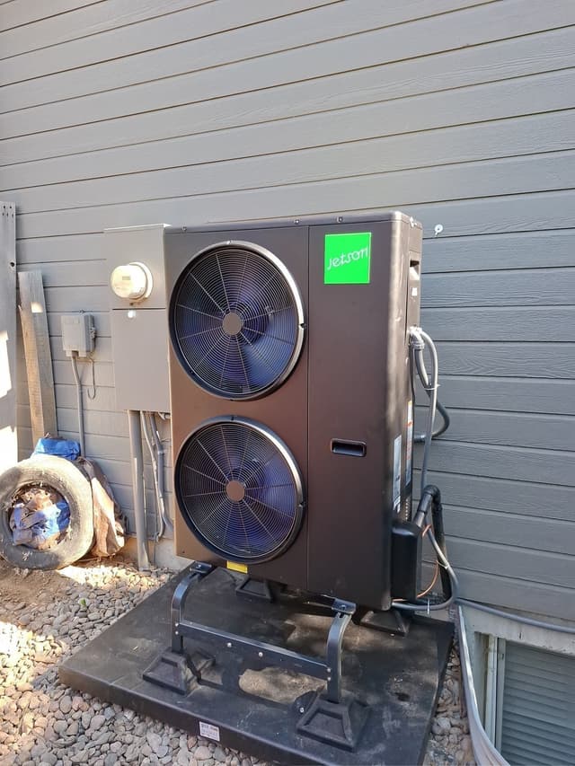 Jetson Air dual-fan 4-ton outdoor heat pump unit on a black metal stand over gravel alongside a gray lap siding house in Commerce City, CO, with refrigerant lines, conduit, disconnect box, and utility meter visible.