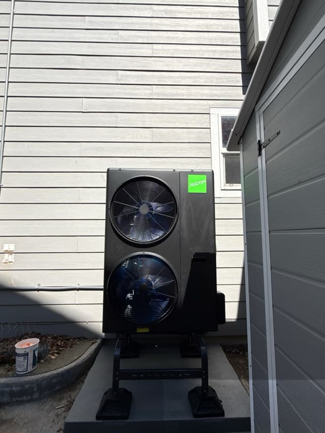 Black Jetson Air dual-fan heat pump condenser mounted on a metal riser stand against gray lap siding in a narrow side yard in Commerce City, CO.