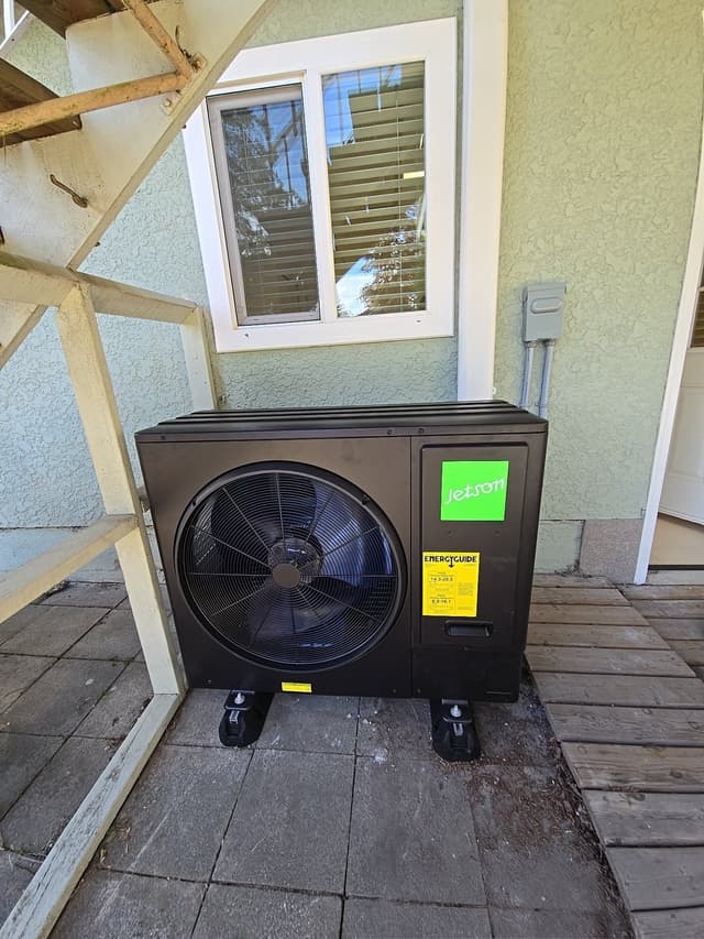 A black Jetson Air 3-ton heat pump installed on anti-vibration feet on a paved patio against a light green stucco exterior wall in Coquitlam, BC.