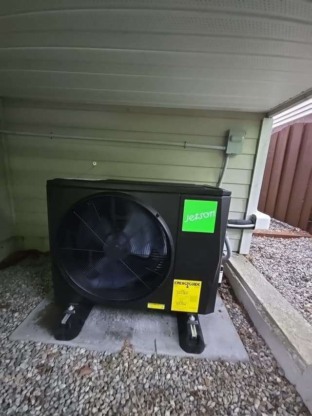 Black Jetson Air 2-ton central ducted heat pump on a concrete pad in a gravel bed, installed under a carport overhang with white soffits, refrigerant lines and electrical disconnect visible against green lap siding, Oxford Heights, Coquitlam, BC.