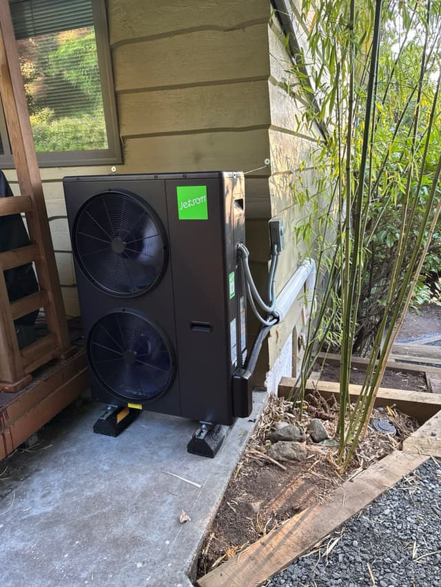 Black Jetson Air 4-ton dual-fan heat pump condenser installed in a corner of a house exterior in Harbour Chines, Coquitlam, with refrigerant lines running along the wall and bamboo stalks to the right.