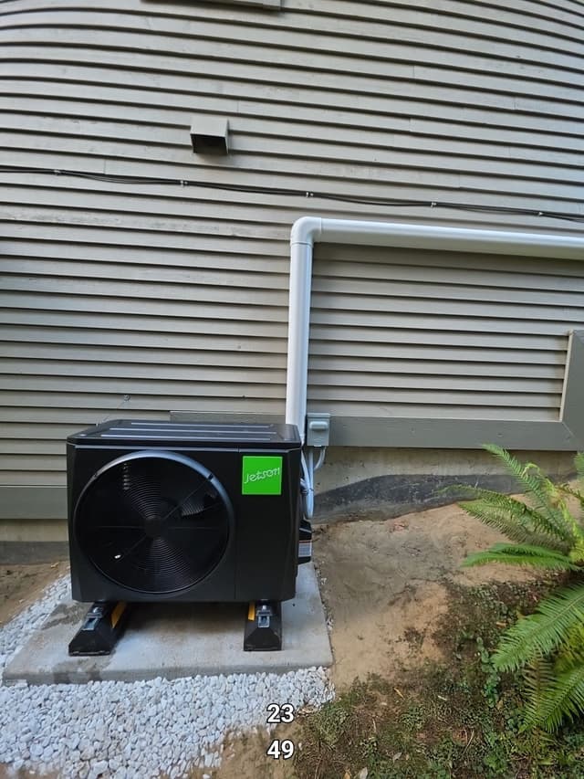 Black Jetson Air outdoor heat pump unit installed against grey horizontal siding at a Westwood Plateau, Coquitlam home, with white gravel surround, grey PVC lineset cover, and a fern visible at lower right.