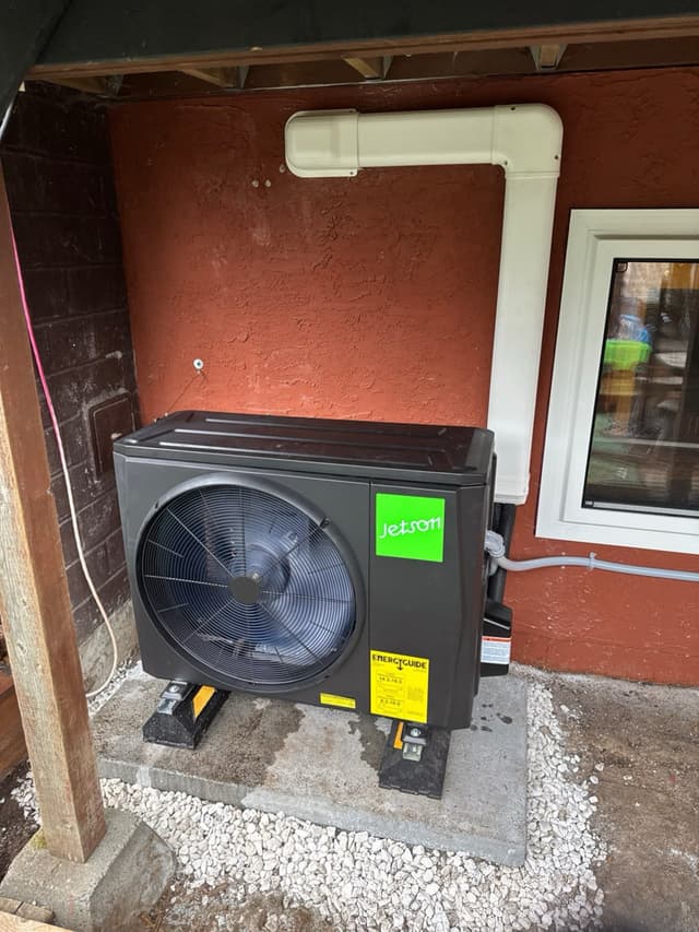 Black Jetson Air 2-ton central ducted heat pump condenser mounted on rubber isolation feet on a gravel and concrete pad against a red stucco wall in River Springs, Coquitlam, BC.