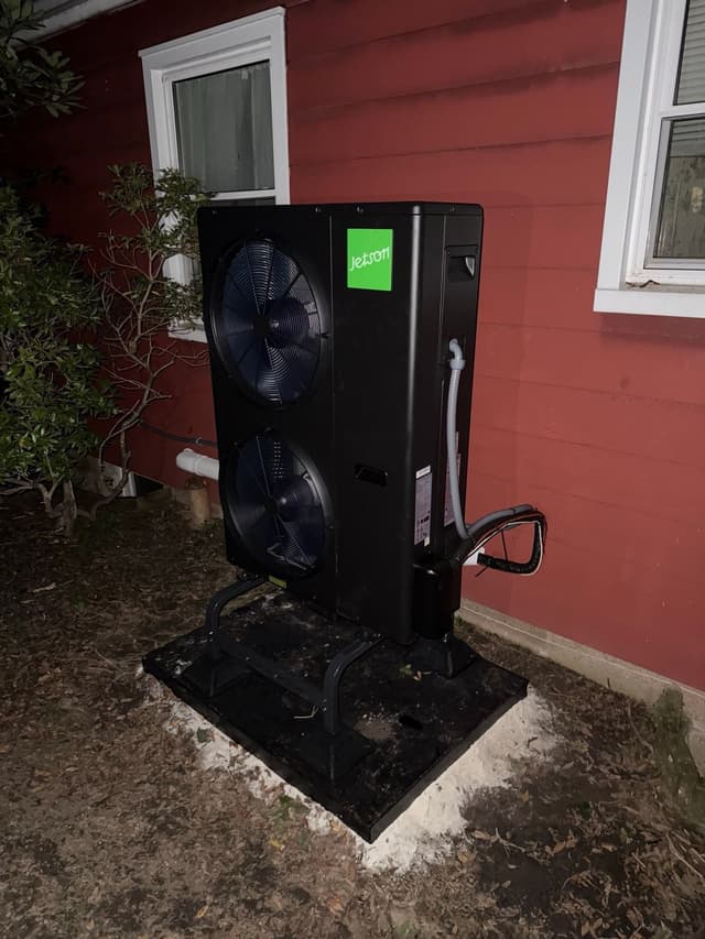 Black Jetson Air 4-ton heat pump installed along the side of a red-sided house in Croton-on-Hudson, NY, photographed at night.