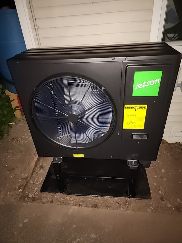 Black Jetson Air 3-ton heat pump condenser on a metal stand against white house siding at night, Upper Village, Croton-on-Hudson, NY.
