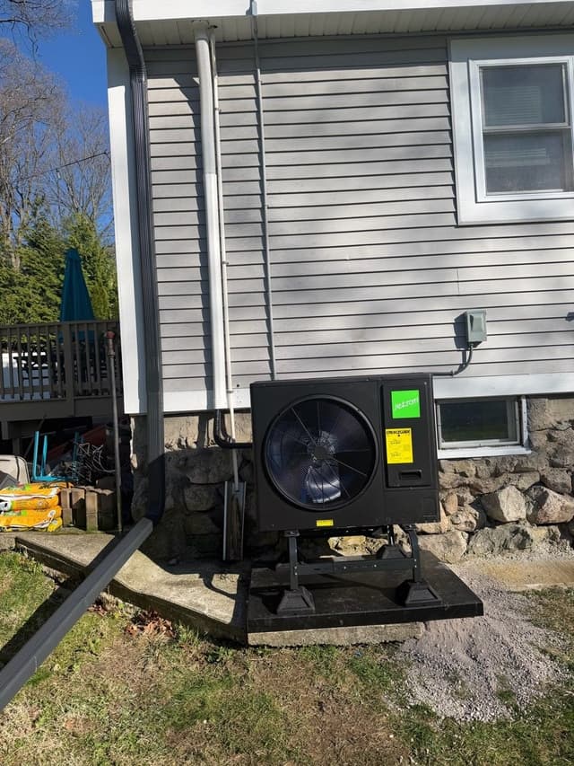 Black Jetson Air 3-ton outdoor heat pump on a stand and concrete pad, with lineset and conduit running up gray vinyl siding on a home in Dedham, MA.