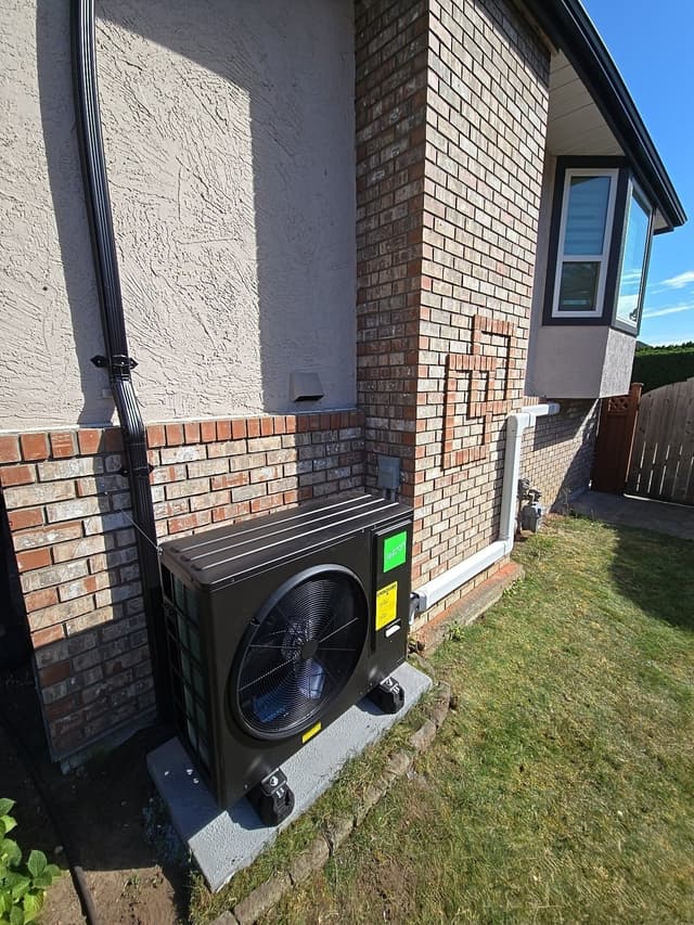 Black Jetson Air 3-ton heat pump condenser on a concrete pad along the side of a brick and stucco home in Delta, BC, with refrigerant lines running up the wall and a clear blue sky in the background.