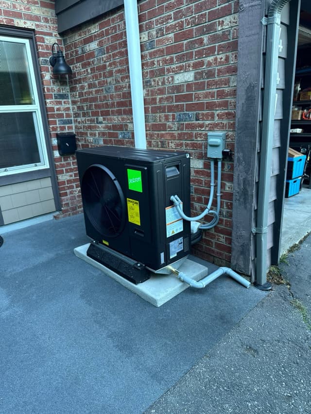 Black Jetson Air 3-ton heat pump condenser on a concrete pad along the brick side wall of a home in Tsawwassen, Delta, BC, with conduit and refrigerant lines running up to a disconnect box.