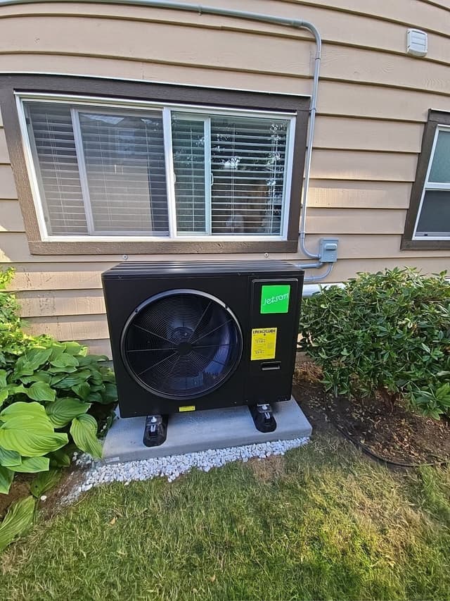 Jetson Air 3-ton heat pump on a concrete pad with vibration-isolating feet along the side of a tan lap siding home in North Delta, Delta BC, flanked by green shrubs, with conduit running up to a wall-mounted electrical disconnect.